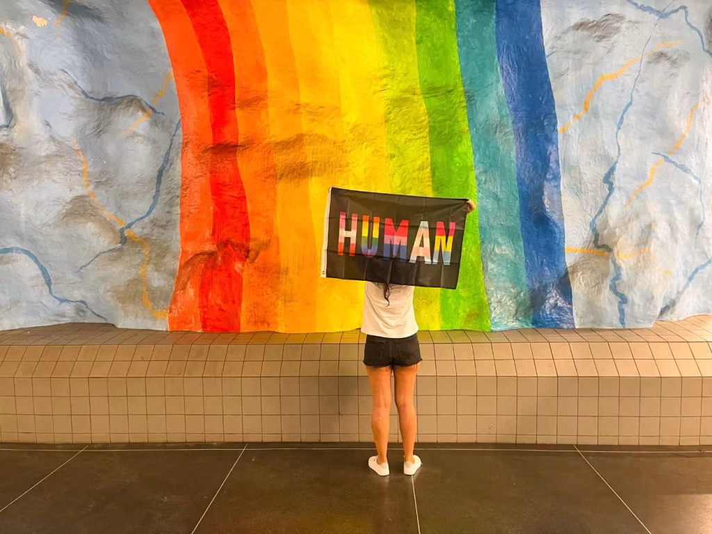 Ragazza con bandiera con su scritto "Human" davanti al dipinto di un arcobaleno